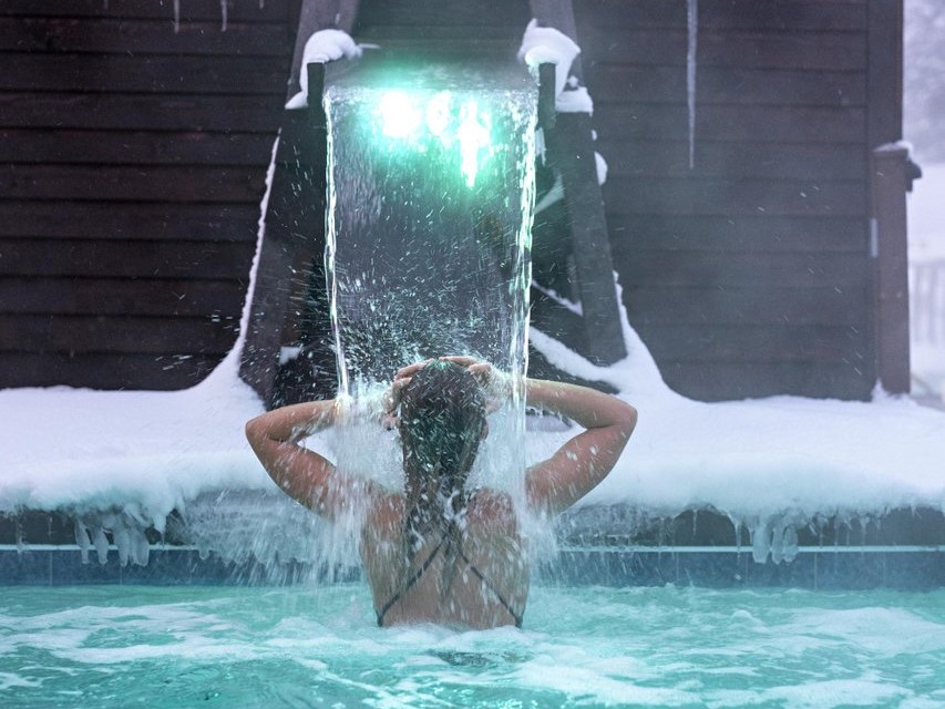 Nordic Spa at Mount Bohemia A woman under a water feature in an outdoor pool during winter.